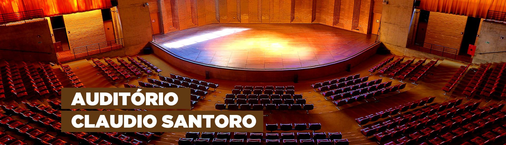 A imagem mostra o Auditório Claudio Santoro que possui uma arquitetura que se assemelha a arena. Há diversas poltronas na cor vermelha, um palco com luzes e vidraças nas quais é possível observar a natureza.