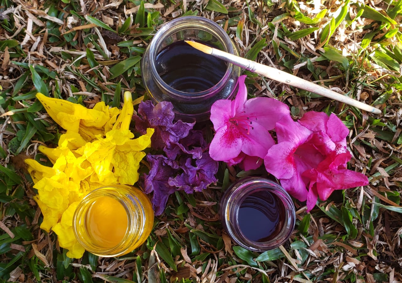 Três potes de vidro com água colorida estão posicionados na grama, ao lado dos potes há pétalas de flores na cor amarela, rosa e roxa. Em um dos potes há um pincel de ponta final.